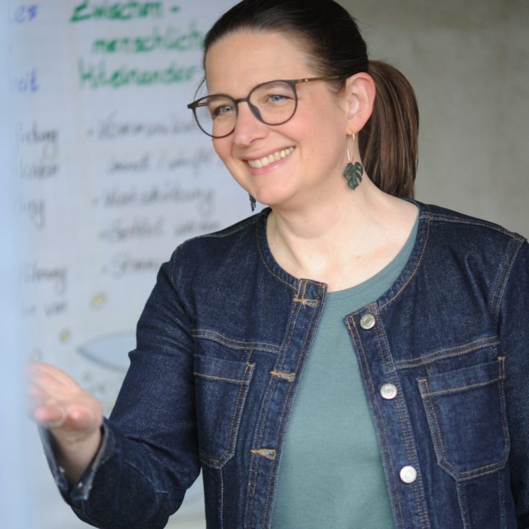 Julia Sittmann Beraterin und Coach Julia Sittmann, Beraterin und Coach, fordert lächelnd eine Person außerhalb des Bildes auf, etwas zu sagen.